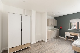 A studio room in 34 Park Street with a large wardrobe, a kitchen unit with cupboard space in the corner of the room and a desk with a chair next to the window against the back wall.